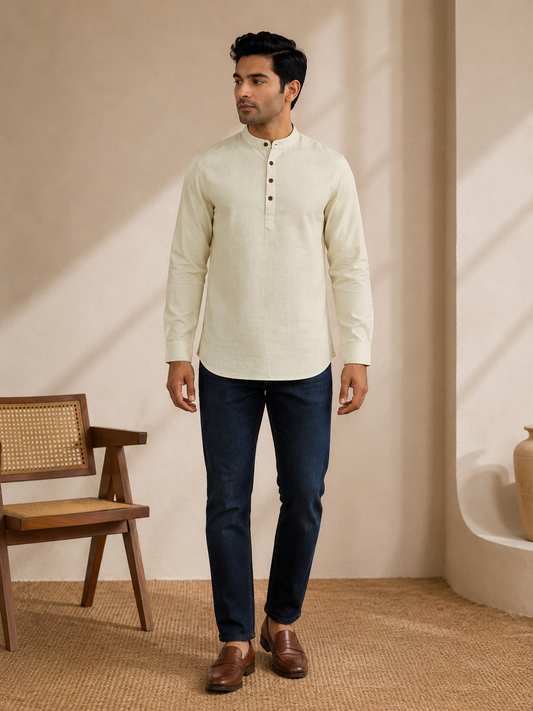 A man wearing a pale lime long-sleeve kurta with a buttoned mandarin collar, paired with dark blue jeans and brown leather shoes, standing in a warm, minimalist room with a wooden chair.