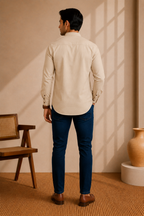 A man viewed from behind wearing a blush beige long-sleeve button-down shirt and dark blue slim-fit jeans, standing in a warm-toned room with a wooden chair and a ceramic vase.