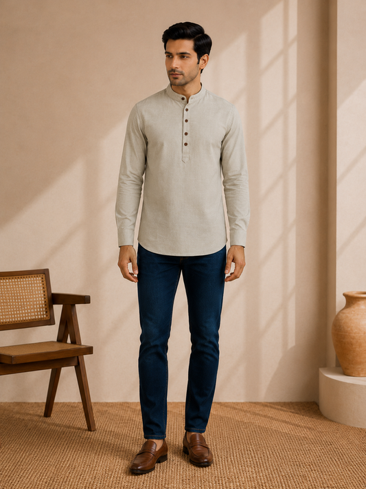 A man wearing a light beige long-sleeve mandarin collar henley shirt, dark blue jeans, and brown leather shoes, standing in a warm-toned room with a wooden chair and a ceramic vase.